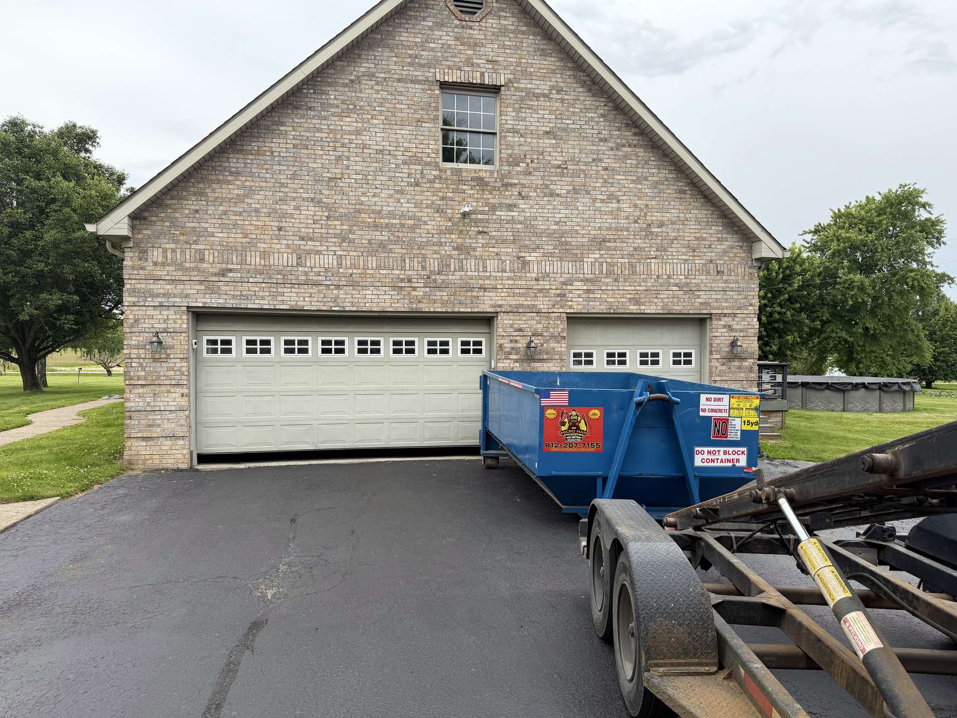 A blue roll-off dumpster set on a driveway in front of a brick garage