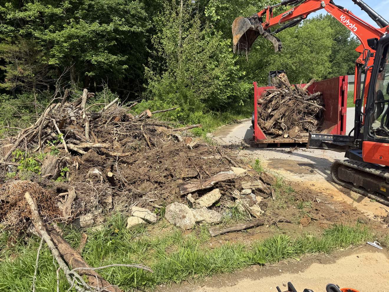 A pile of brush and debris beside a red roll-off dumpster, with an excavator loading it