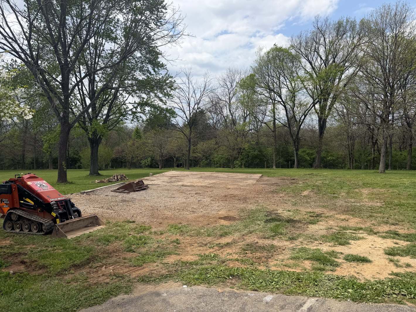 A large cleared and graveled pad in a grassy field, a stand-on loader parked at the edge