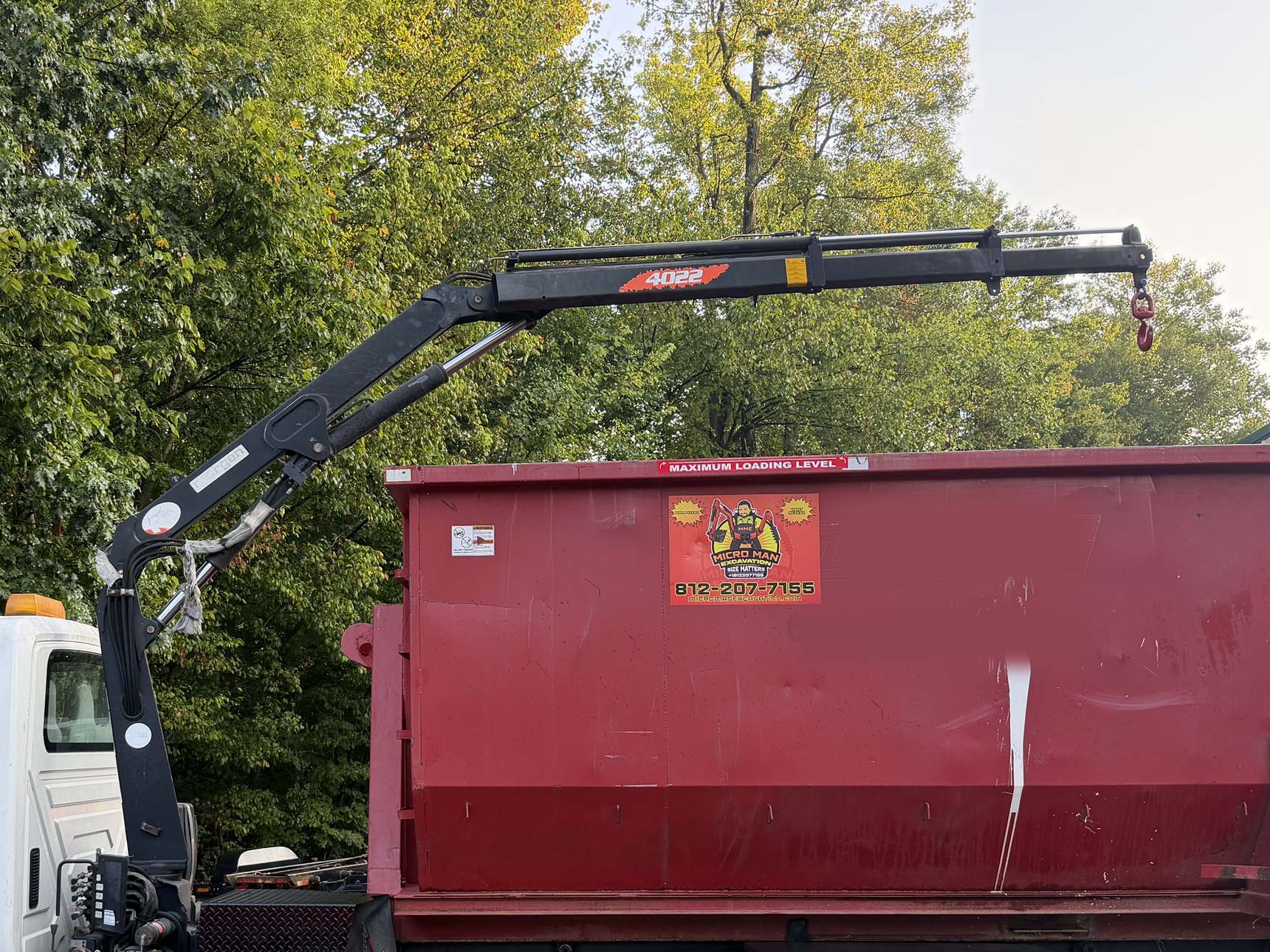 The crane on the Micro Man roll-off truck, its boom folded over a red roll-off dumpster