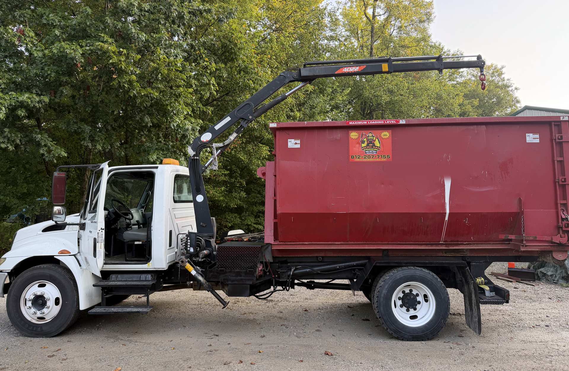 The white Micro Man roll-off truck with its crane folded over a red roll-off dumpster