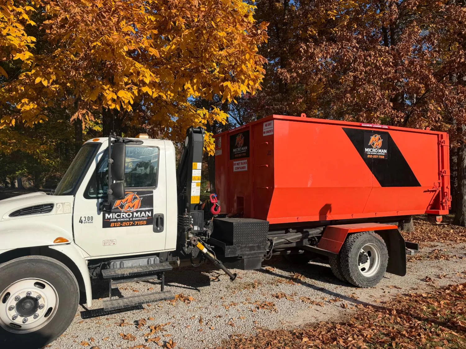 White Micro Man crane truck carrying an orange Micro Man roll-off dumpster on a gravel lot under yellow and rust colored fall trees