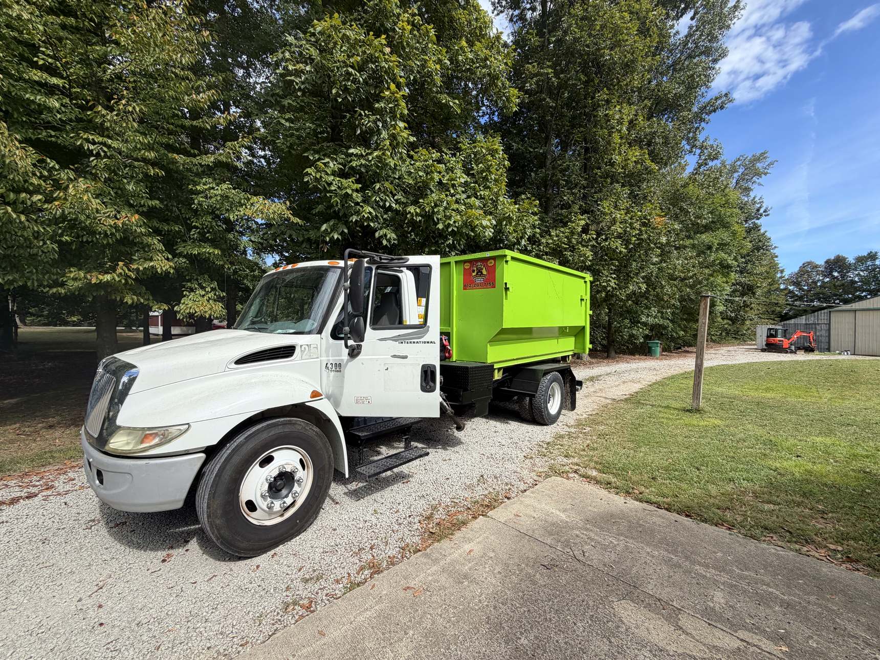 The white Micro Man roll-off truck carrying a green dumpster down a gravel driveway under tall trees