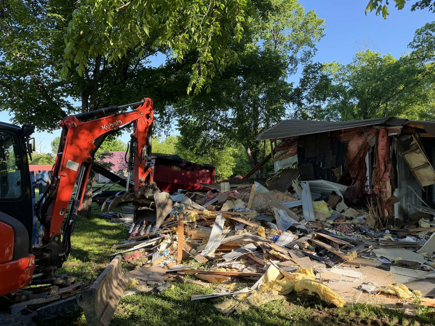 A mini excavator tearing down a small house, a red roll-off waiting behind
