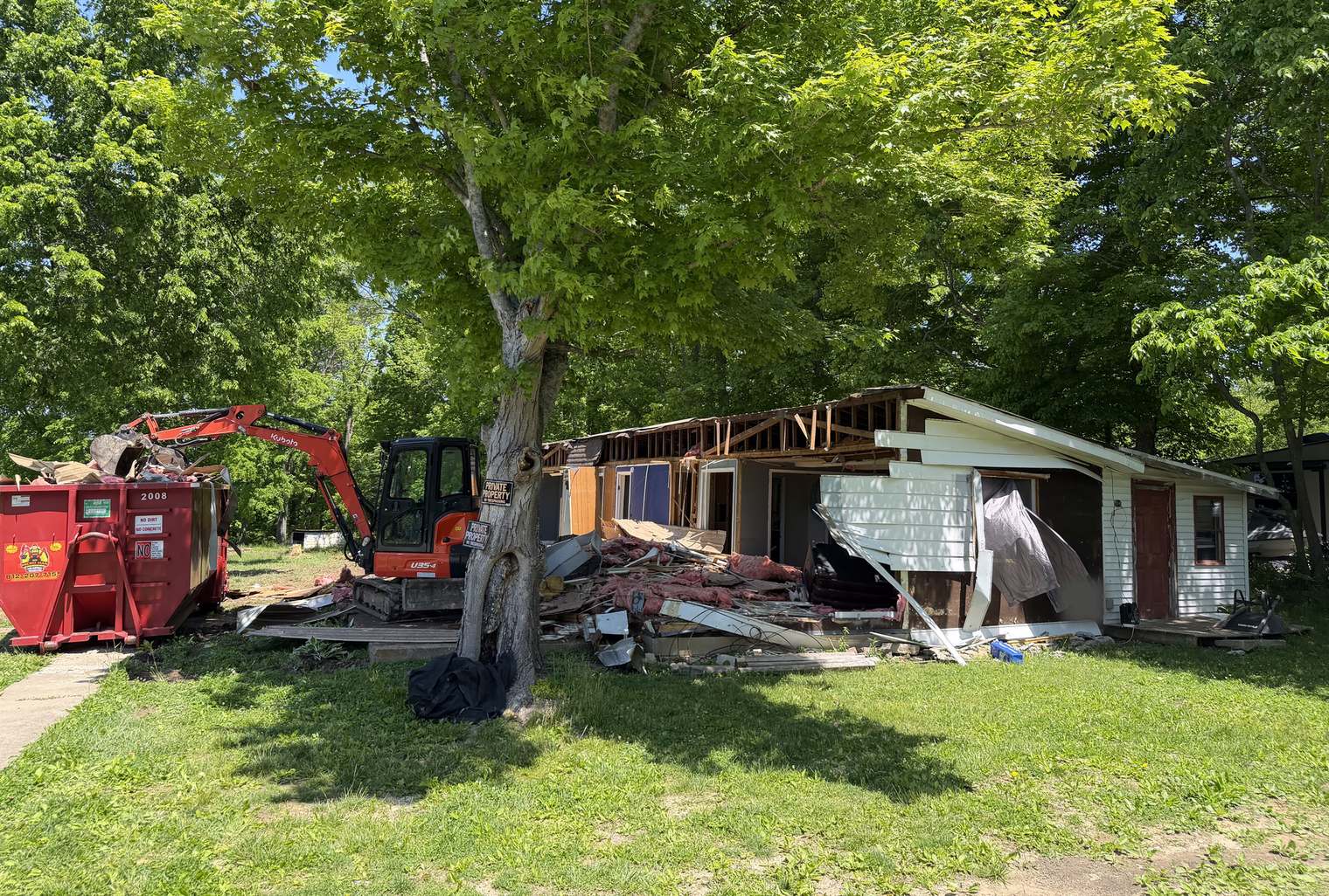 A mini excavator tearing into a small single-story house under shade trees, a red Micro Man roll-off beside it