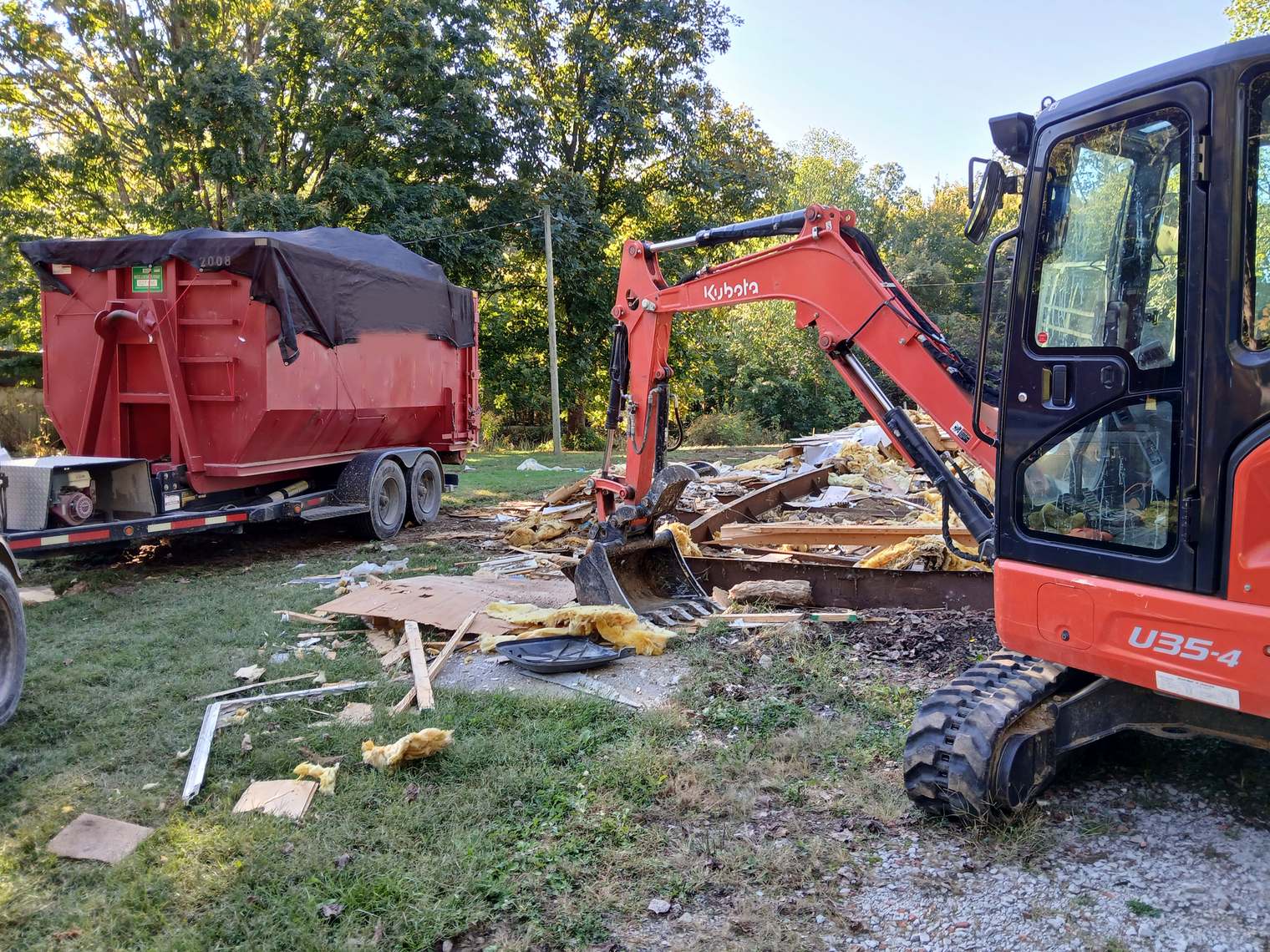 A mini excavator loading teardown debris into a red roll-off on its trailer