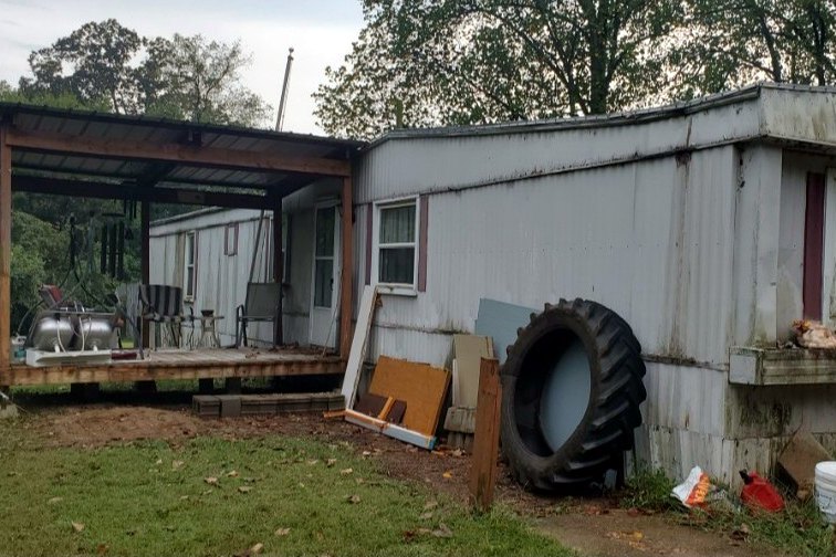 An old mobile home with a covered porch, a tire and debris beside it, before the teardown
