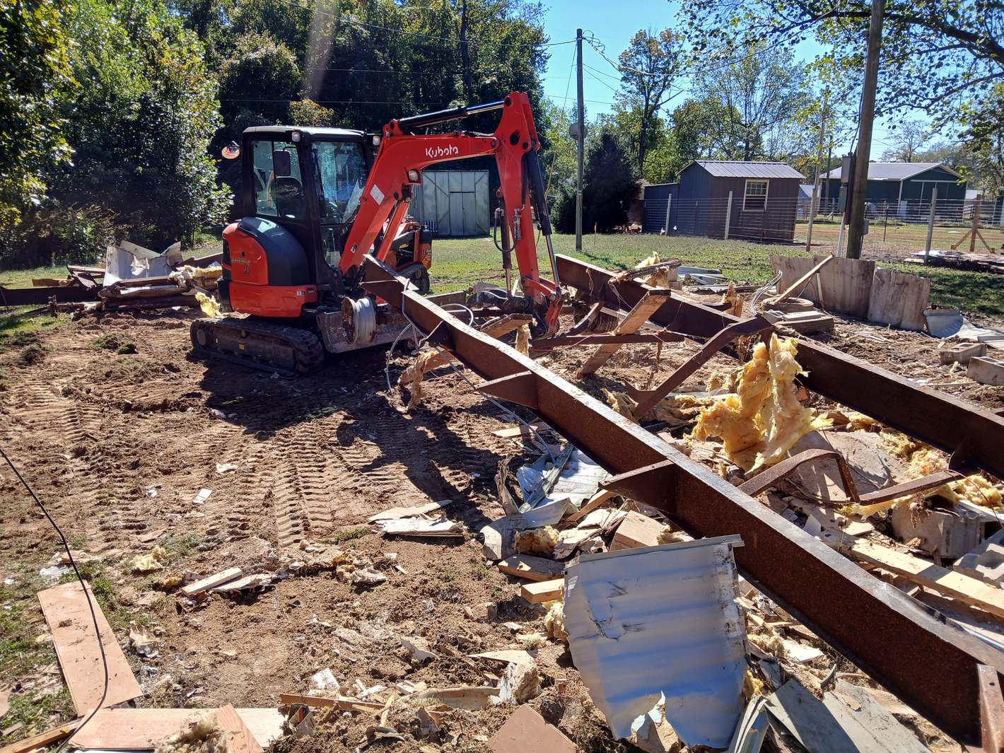 An orange mini excavator beside the steel frame of a mobile home it has torn down, insulation and siding across the lot