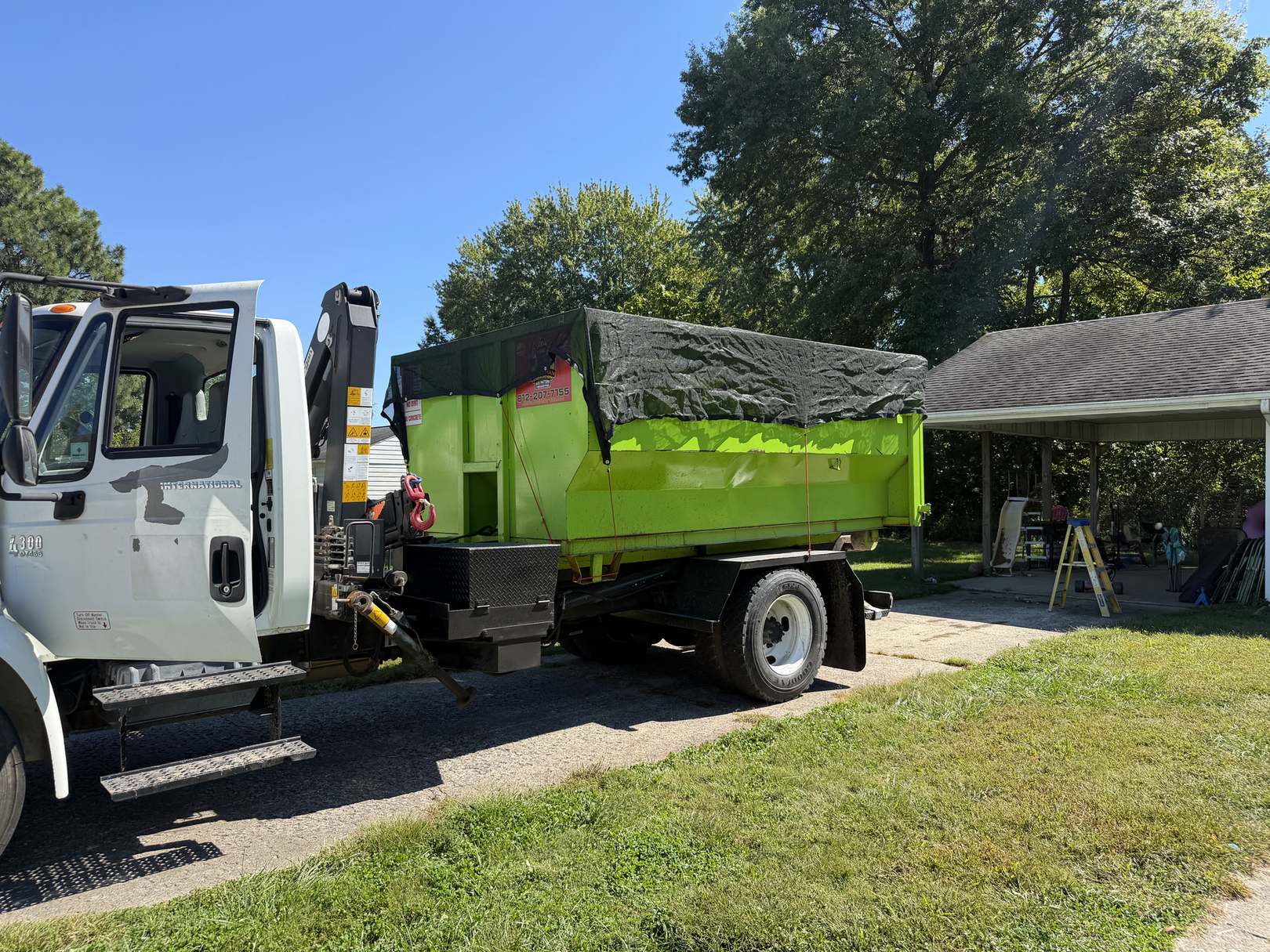 The Micro Man roll-off truck carrying a tarped green dumpster beside a carport