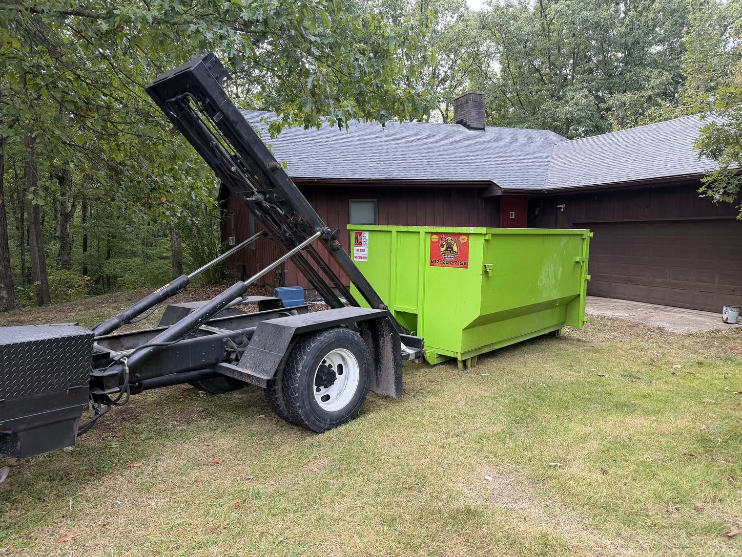 A green Micro Man roll-off set down on a lawn beside a garage, the roll-off truck arm still raised