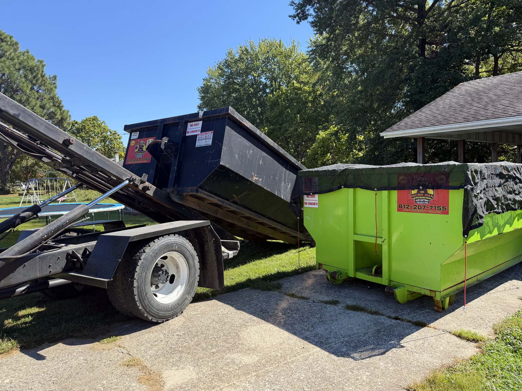 A Micro Man roll-off truck setting down a black dumpster beside a tarped green one on a driveway