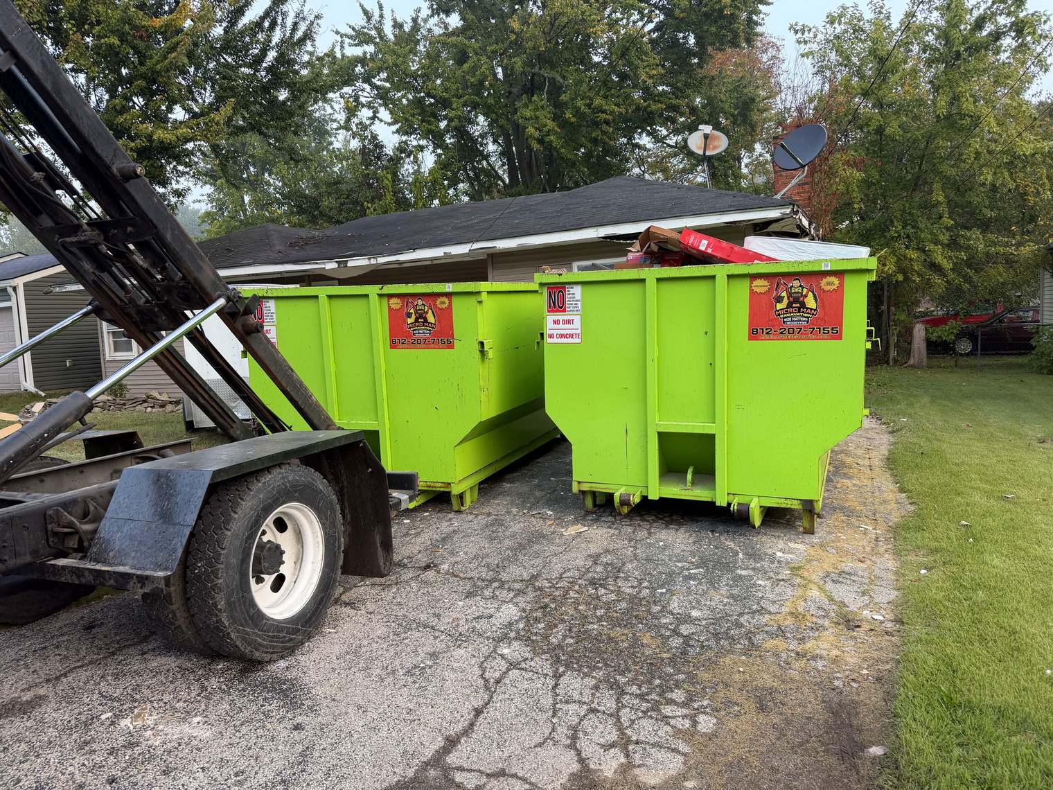 Two green Micro Man roll-off dumpsters side by side on a driveway, one loaded with boxes and a mattress