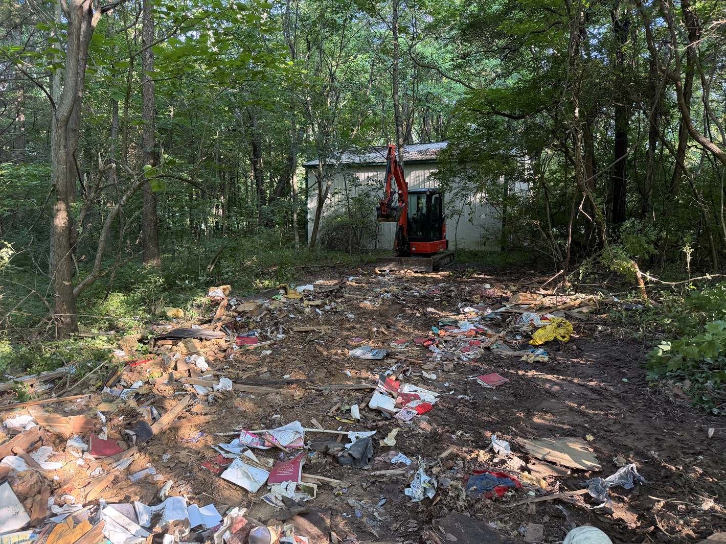 A mini excavator clearing years of scattered trash and debris from a wooded lot